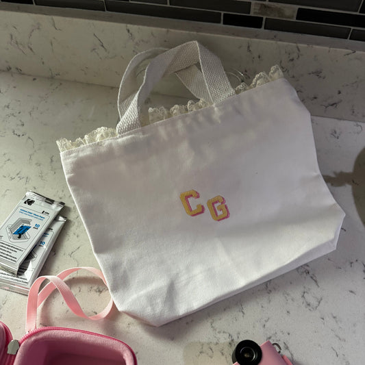 White lace bag with 'CG' initials, pink camera, and pink container on a marble surface.