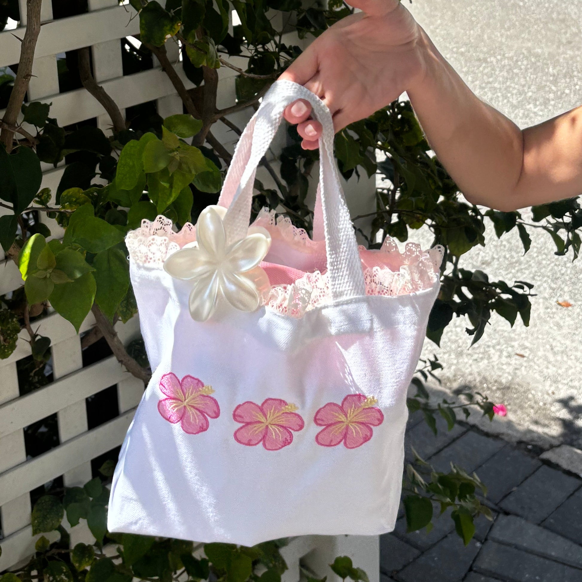Person holding a white bag with pink floral designs near a white lattice fence and green plants.