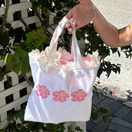 Person holding a white bag with pink floral designs near a white lattice fence and green plants.