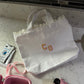 White lace bag with 'CG' initials, pink camera, and pink container on a marble surface.